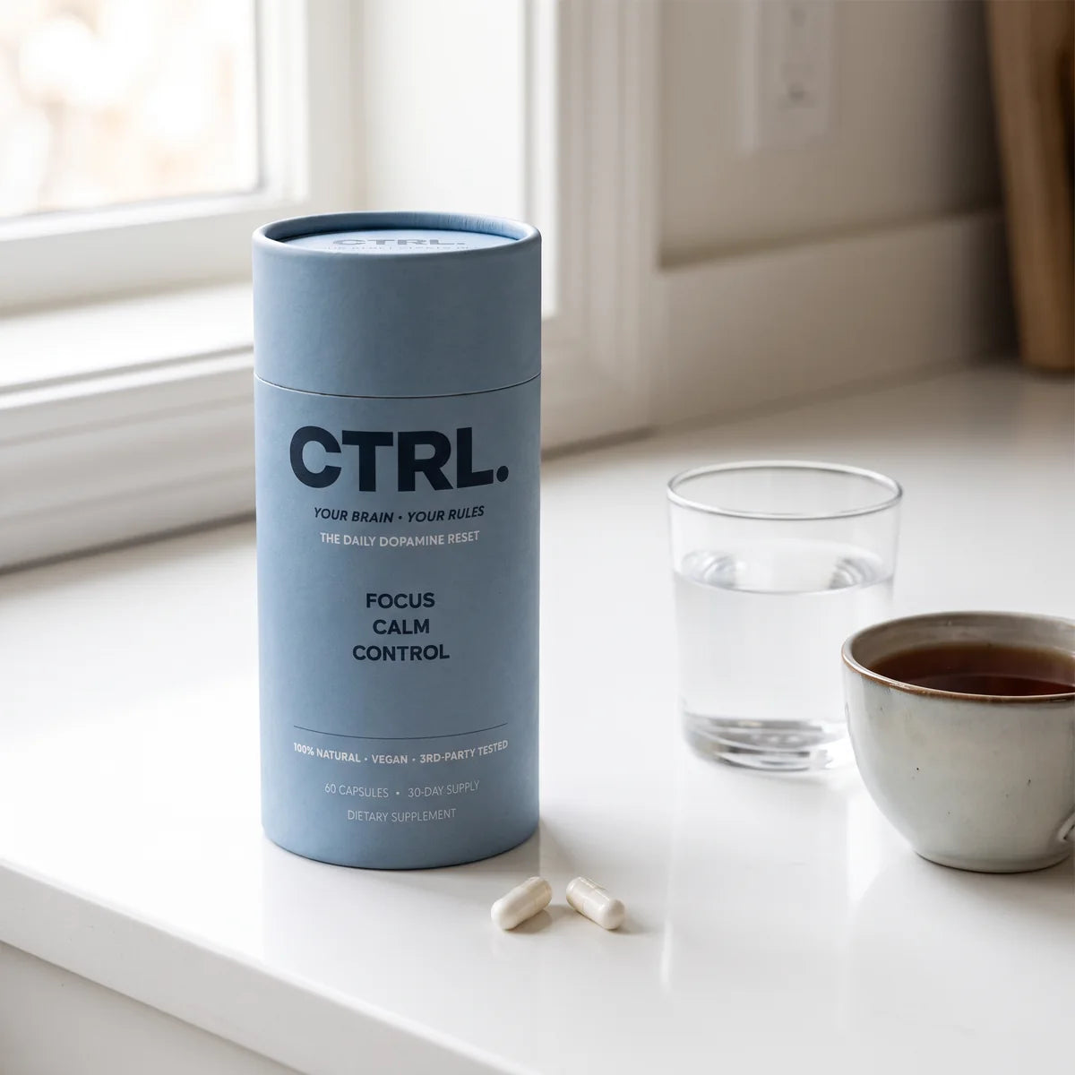 The CTRL tube and two capsules on a bright kitchen counter next to a glass of water and a cup of tea