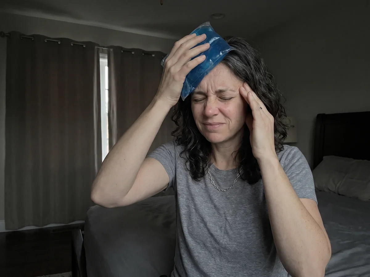 Diane sitting on the edge of her bed in a dim bedroom with the curtains drawn, pressing an ice pack to her forehead, eyes squeezed shut in pain