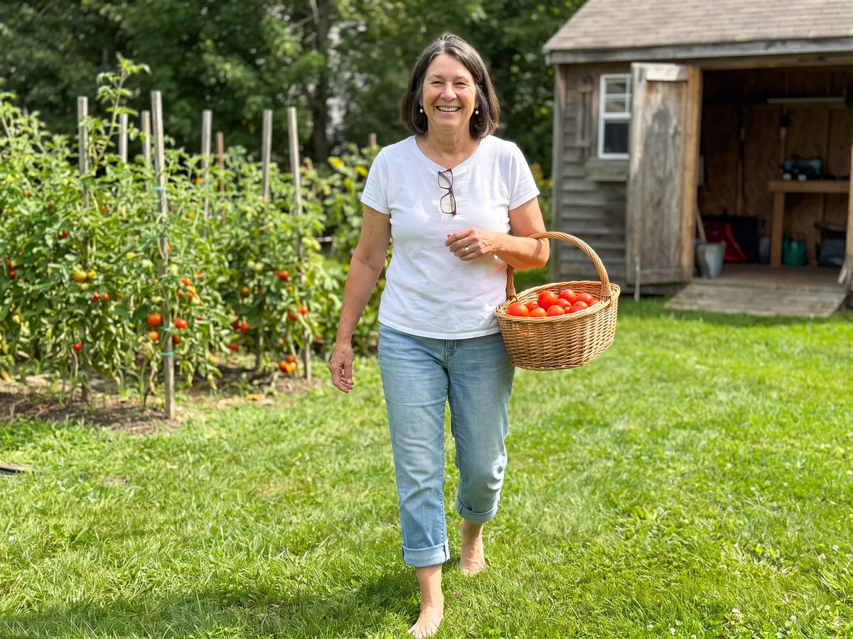 Carol walking barefoot across the green lawn of her vegetable garden on a sunny morning, smiling, with a basket of tomatoes on her arm
