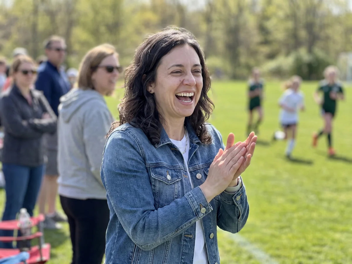 Diane cheering on the sideline of her daughter's soccer game on a bright sunny day, no sunglasses