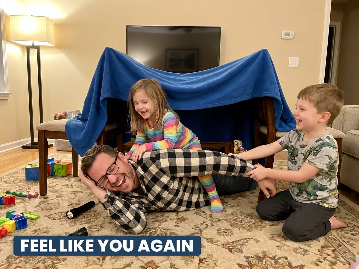 A dad laughing on the living room floor with his two kids in a blanket fort